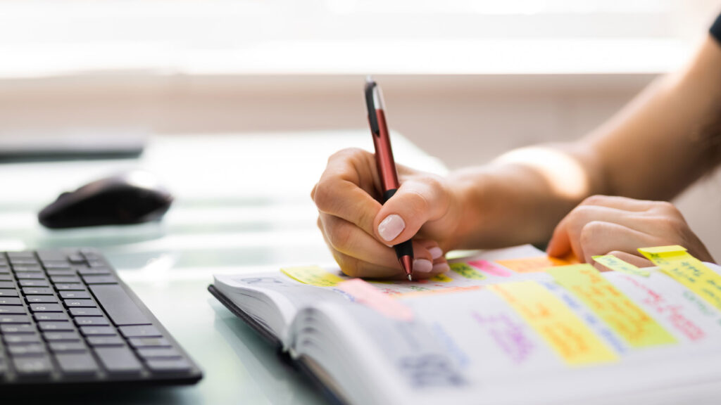 Person writing in a color-coded planner with sticky notes, preparing an experiential marketing event timeline at a desk with keyboard and mouse.