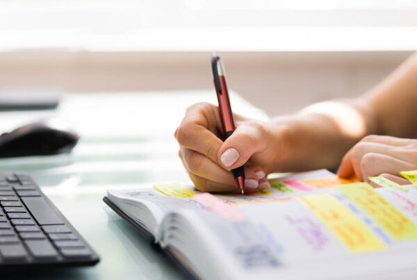Person writing in a color-coded planner with sticky notes, preparing an experiential marketing event timeline at a desk with keyboard and mouse.