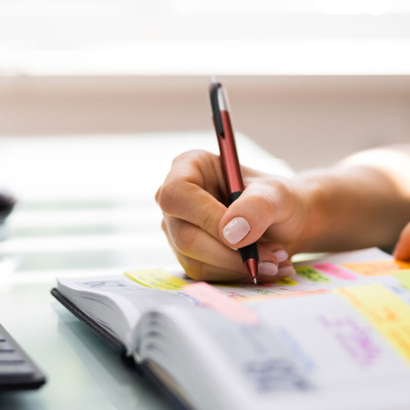 Person writing in a color-coded planner with sticky notes, preparing an experiential marketing event timeline at a desk with keyboard and mouse.