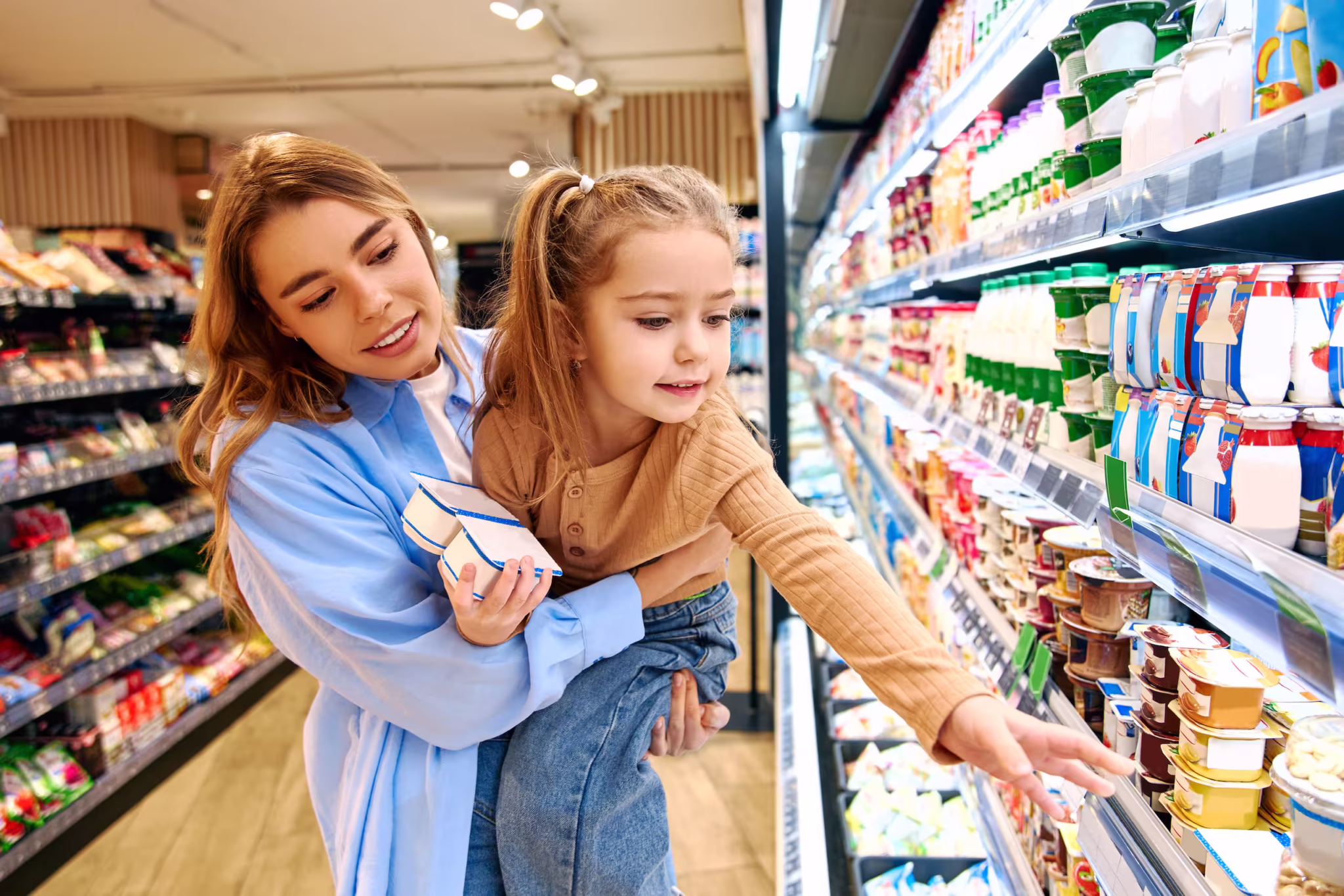 Parent and child choosing yogurt in a grocery dairy aisle, illustrating consumer packaged goods decisions at the shelf.